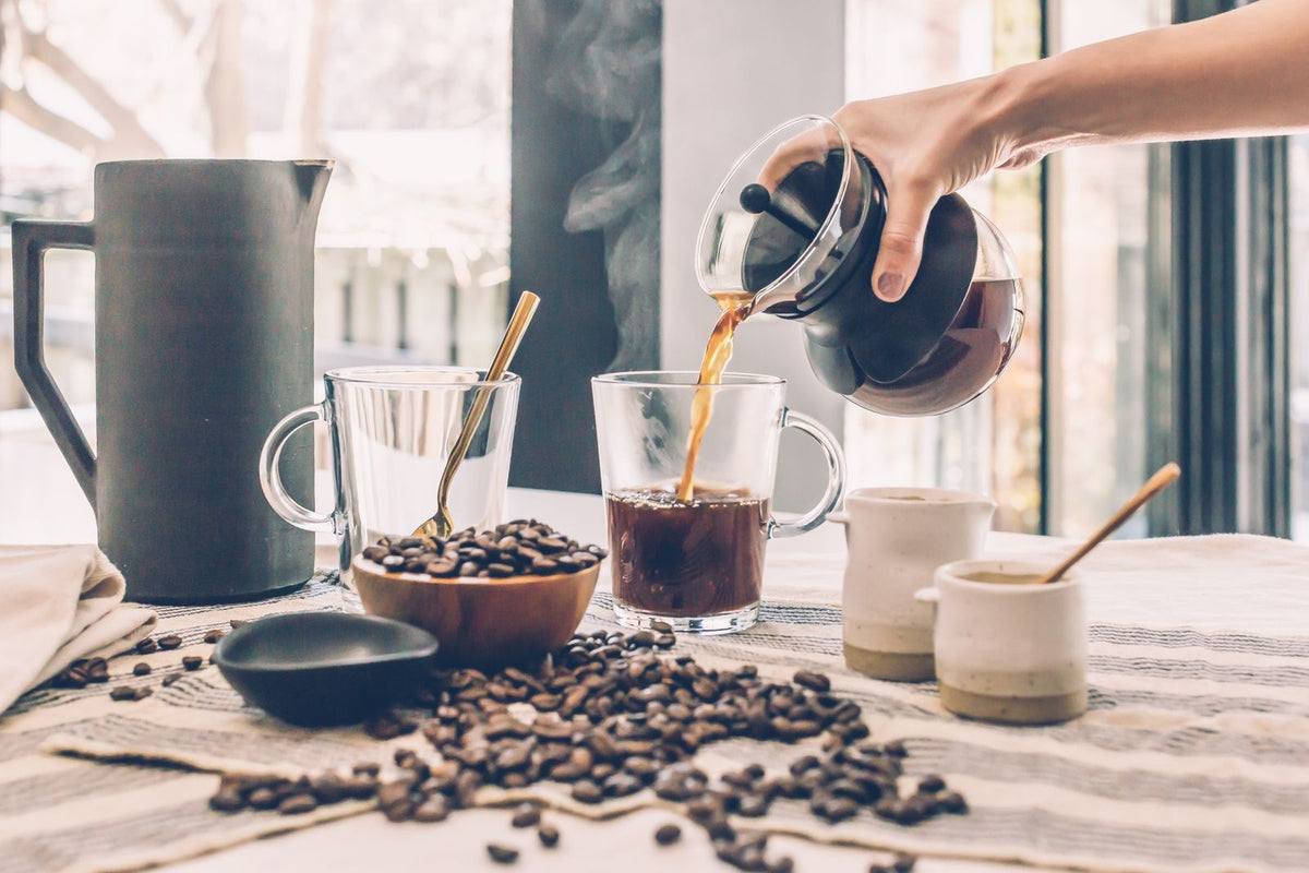 Hands pouring morning coffee into glass mugs on a sunlit table, a quiet routine scene that pairs well with everyday wellness supplements