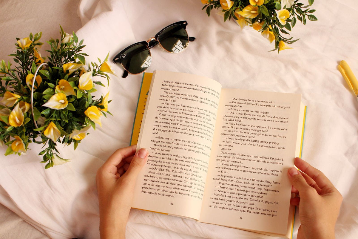 Hands holding an open book on a bright surface with small flowers nearby, a calm reading setup supporting everyday cognitive lifestyle habits
